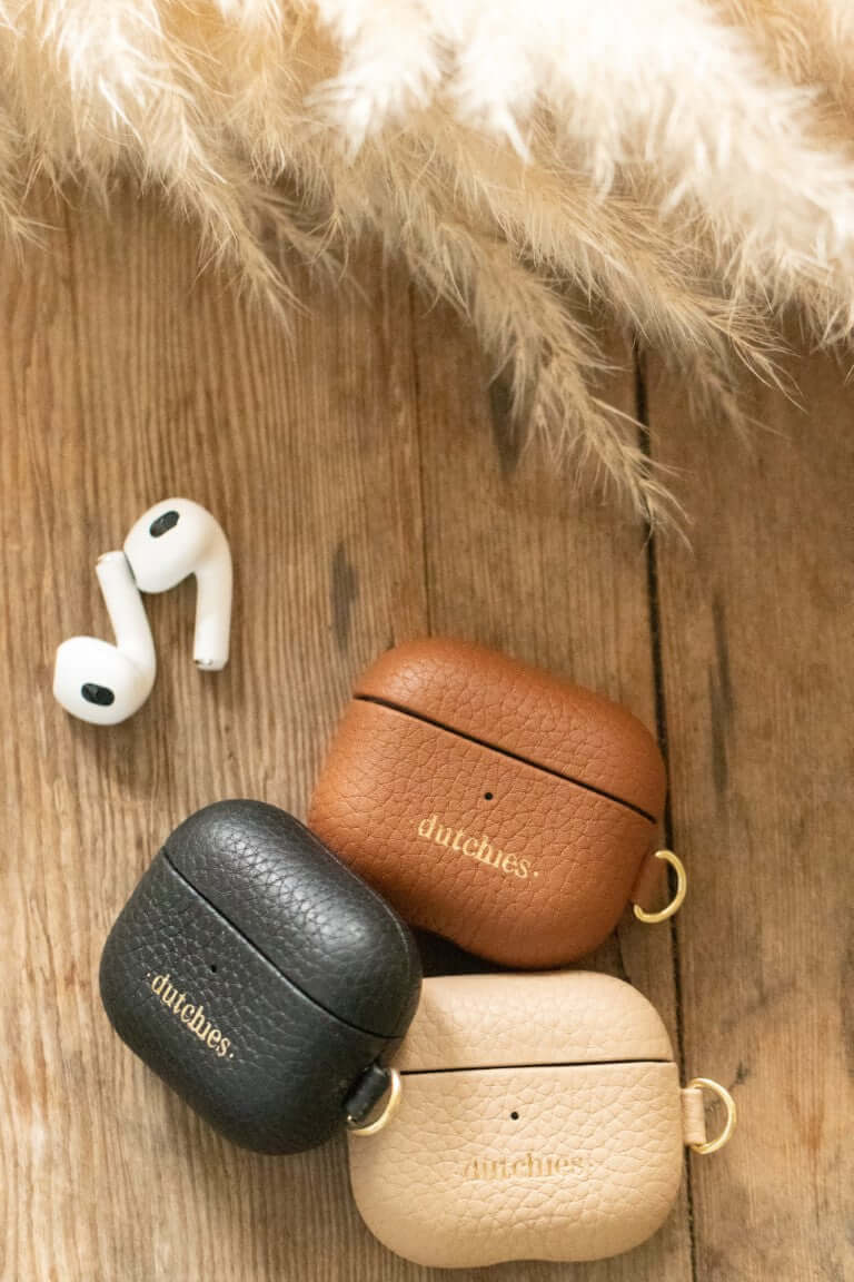 Airpod Cases in brown, beige, and black leather from Dutchies on a wooden surface with AirPods.
