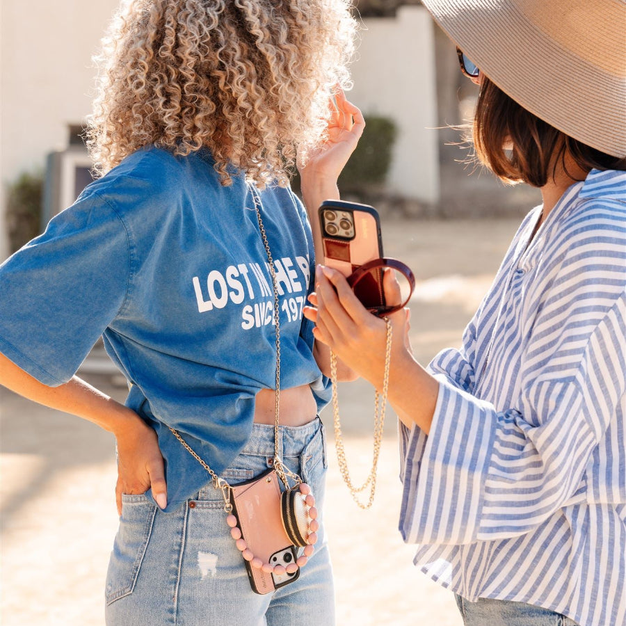 Two women showing off phone charms in sunny outdoor setting, wearing stylish outfits and accessories.