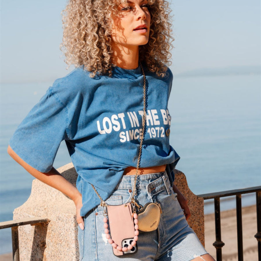 Model wearing a blue shirt and shorts, showcasing a pink phone charm attached to a phone case.