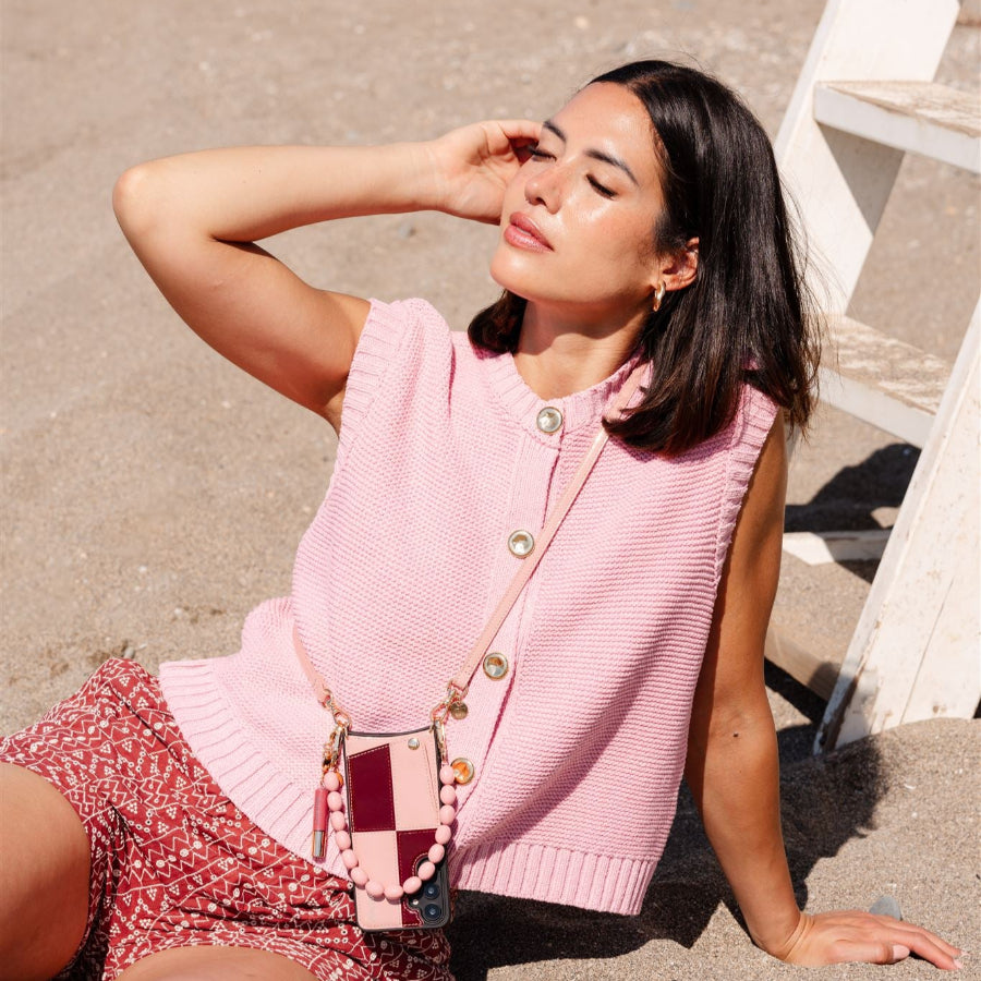 Model wearing a pink sleeveless sweater and patterned shorts, showcasing a pink phone charm on the beach.