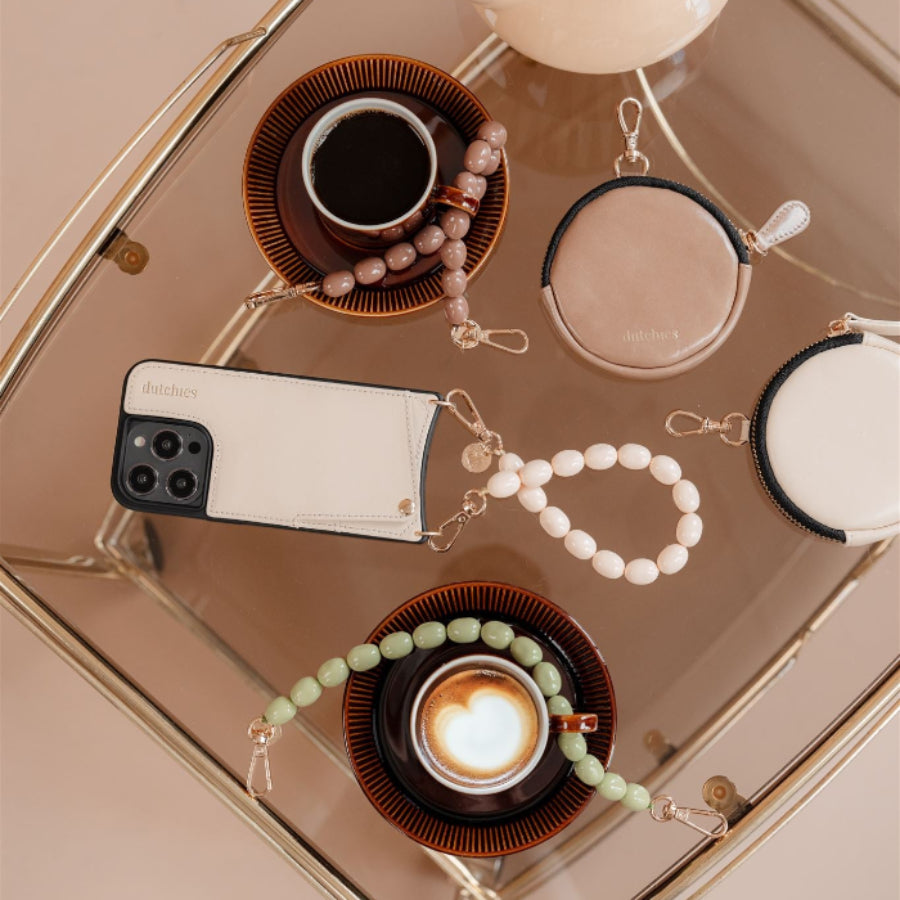 Flat lay of beaded phone charm and stylish phone case with coffee cups on a chic table.