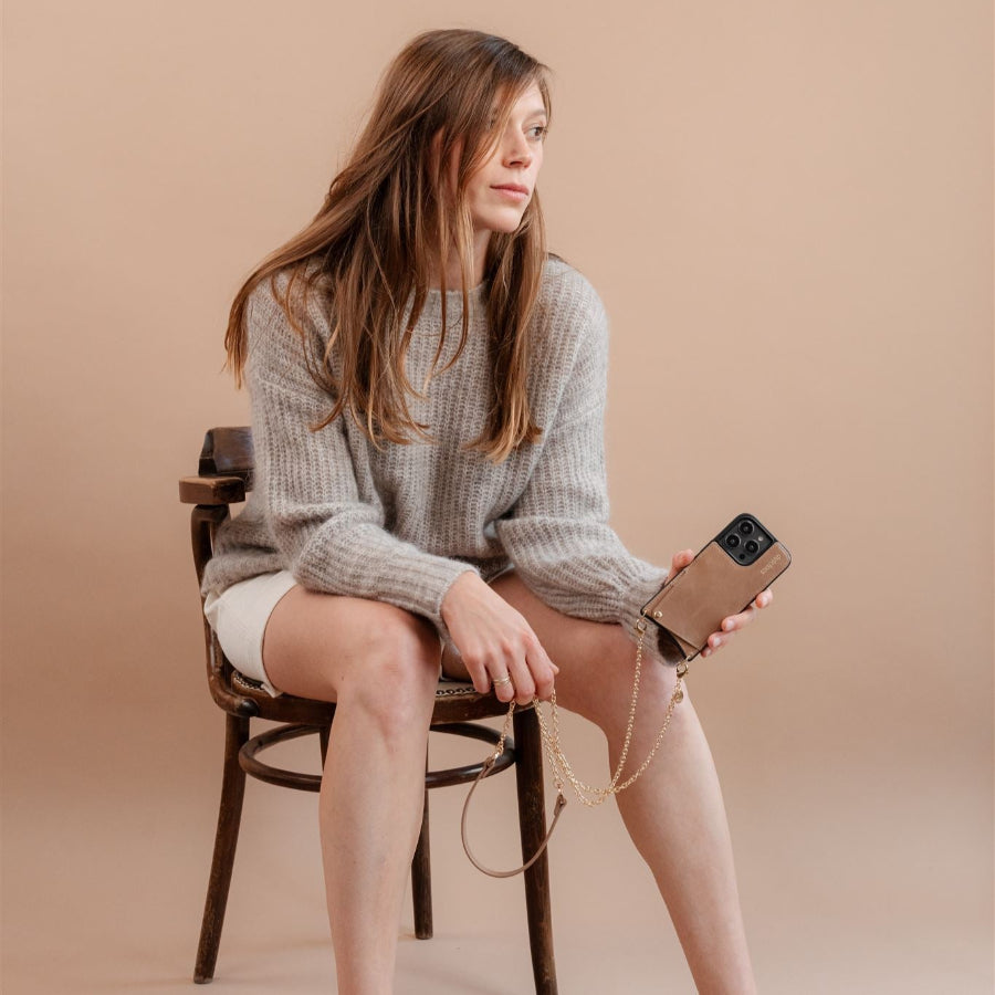 Woman sitting on a chair holding an iPhone Case | Mocha with a stylish strap, showcasing its chic design.