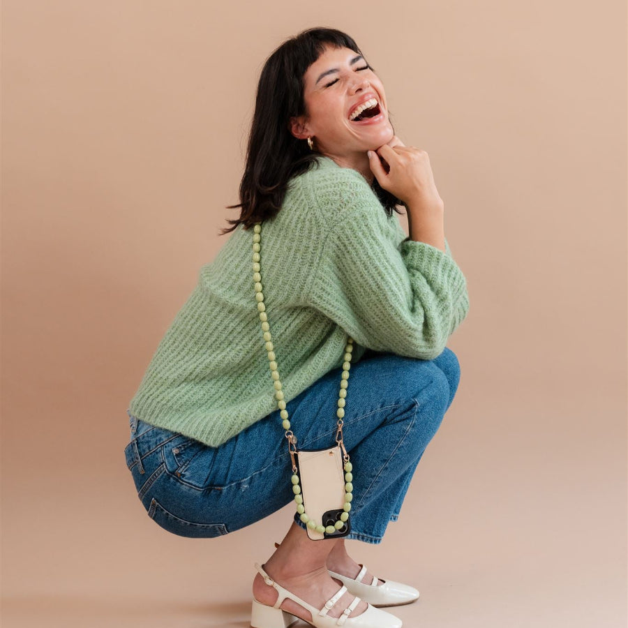 Smiling woman in green sweater squatting with a beaded phone charm hanging from her phone.