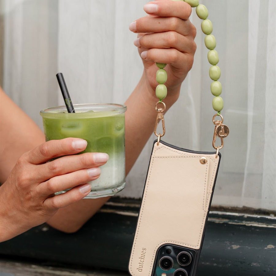 Beaded phone charm in green held by a hand next to a refreshing drink, blending style and functionality.