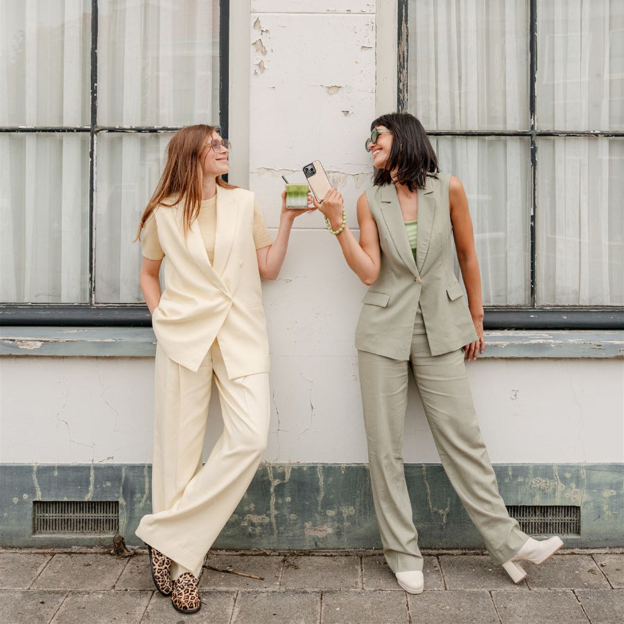 Two women in stylish suits holding drinks and showing off their phone charms in an urban setting.