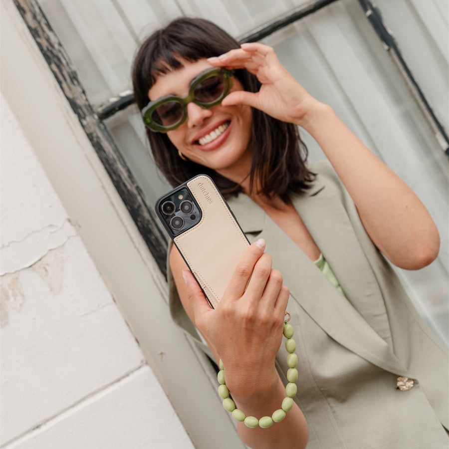 Woman holding a phone with a beaded green charm, wearing stylish green sunglasses and a beige outfit, outdoors.