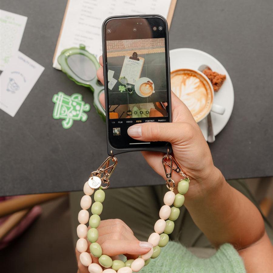 A hand holding a smartphone with a beaded charm, capturing a coffee scene at a café.