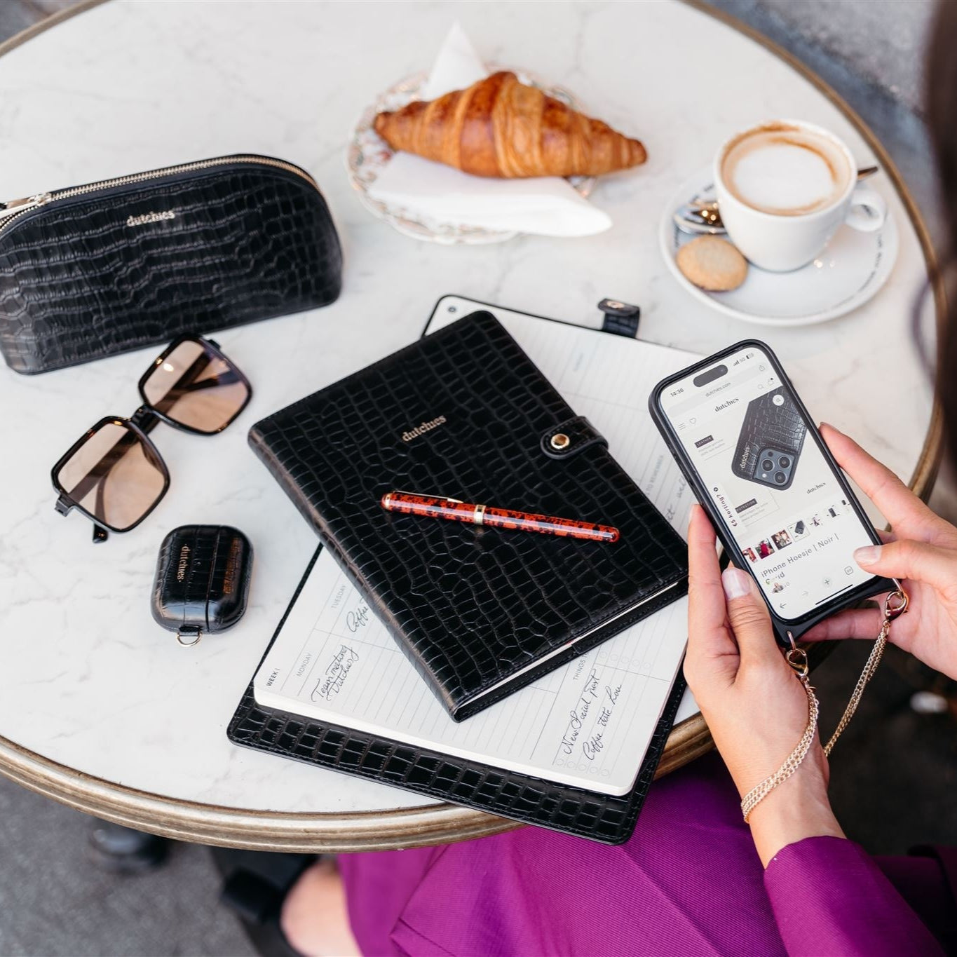 Stylish black leather notebook with gold details on a cafe table, beside sunglasses, smartphone, and coffee.