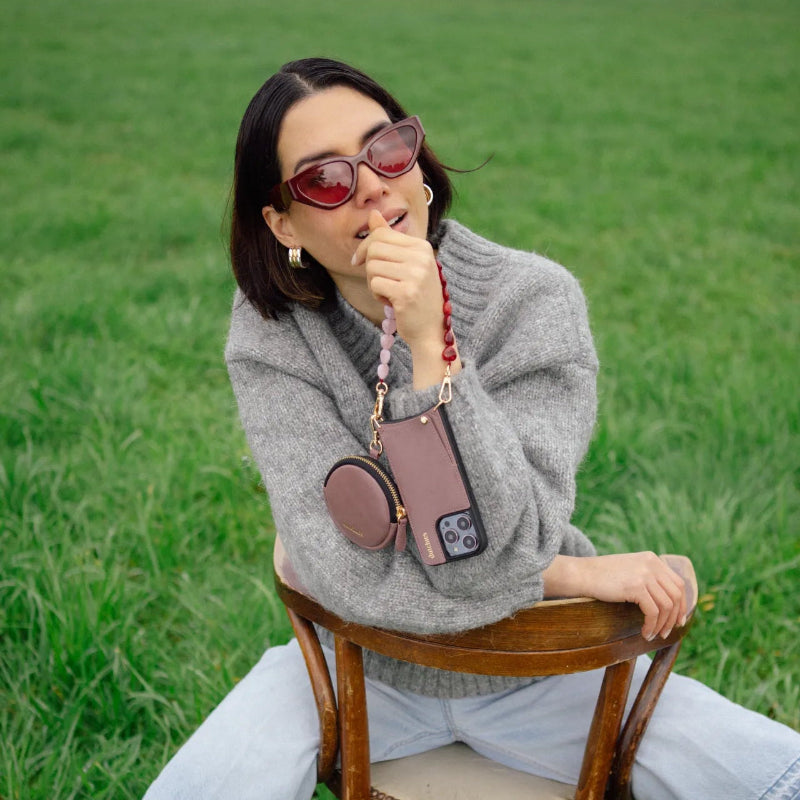 Woman sitting on a chair wearing stylish sunglasses, showcasing the Mini Phone Bag Emma | Mauve clipped to her phone.