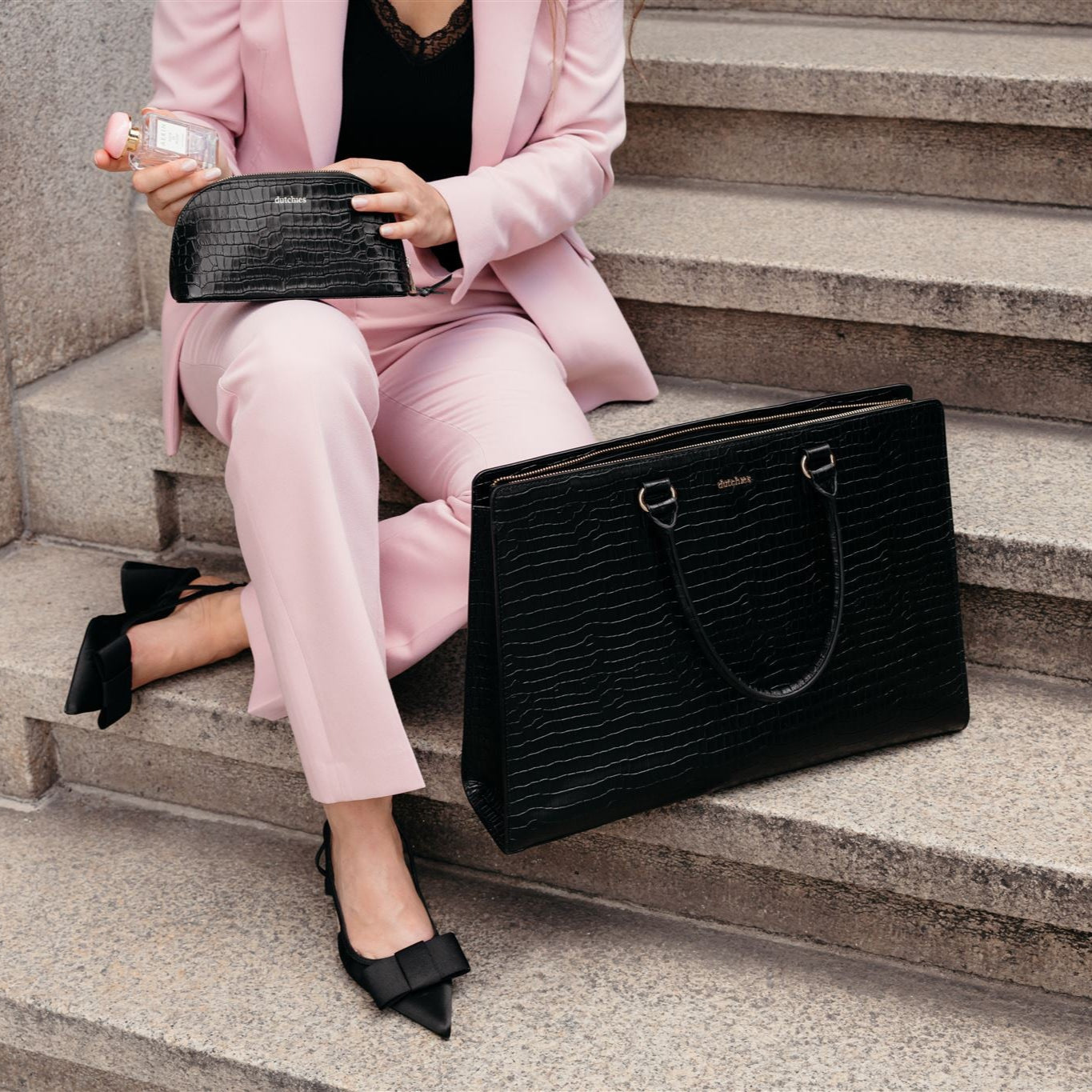 Stylish woman in pink suit with black textured bags sitting on stairs.