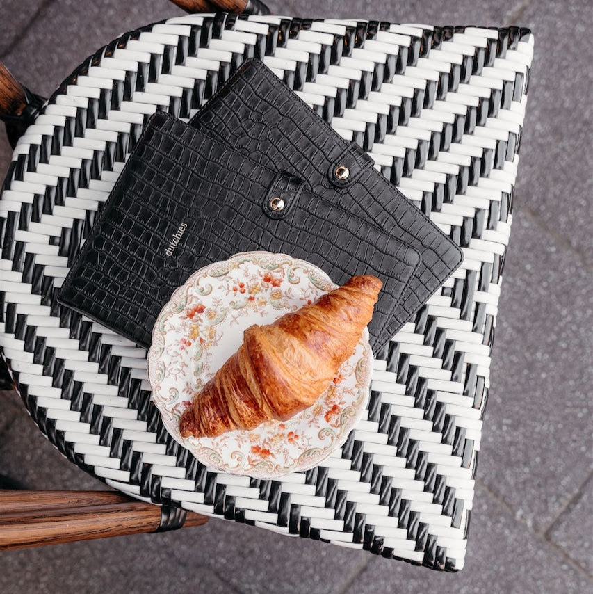 Black leather notebook and croissant on a stylish patterned table, perfect for a chic work or café setting.