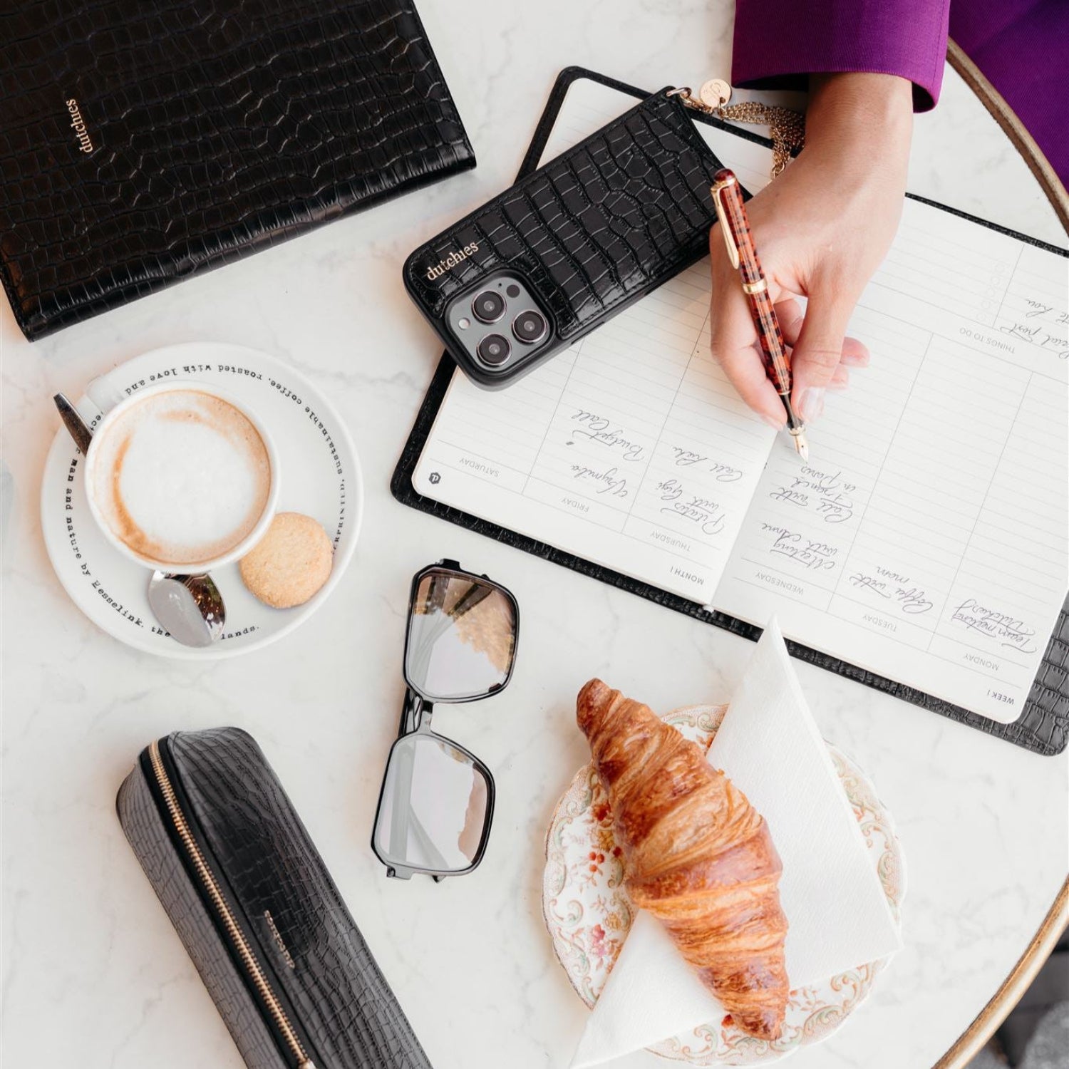 Stylish black leather notebook with gold details, coffee, croissant, and sunglasses on a marble table.