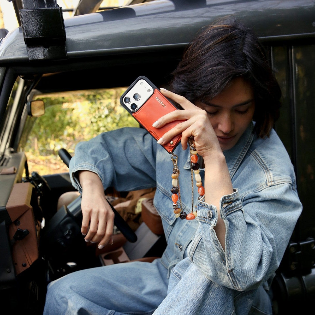 Woman in a denim jacket holding an iPhone Case | Orange with a strap inside a car.
