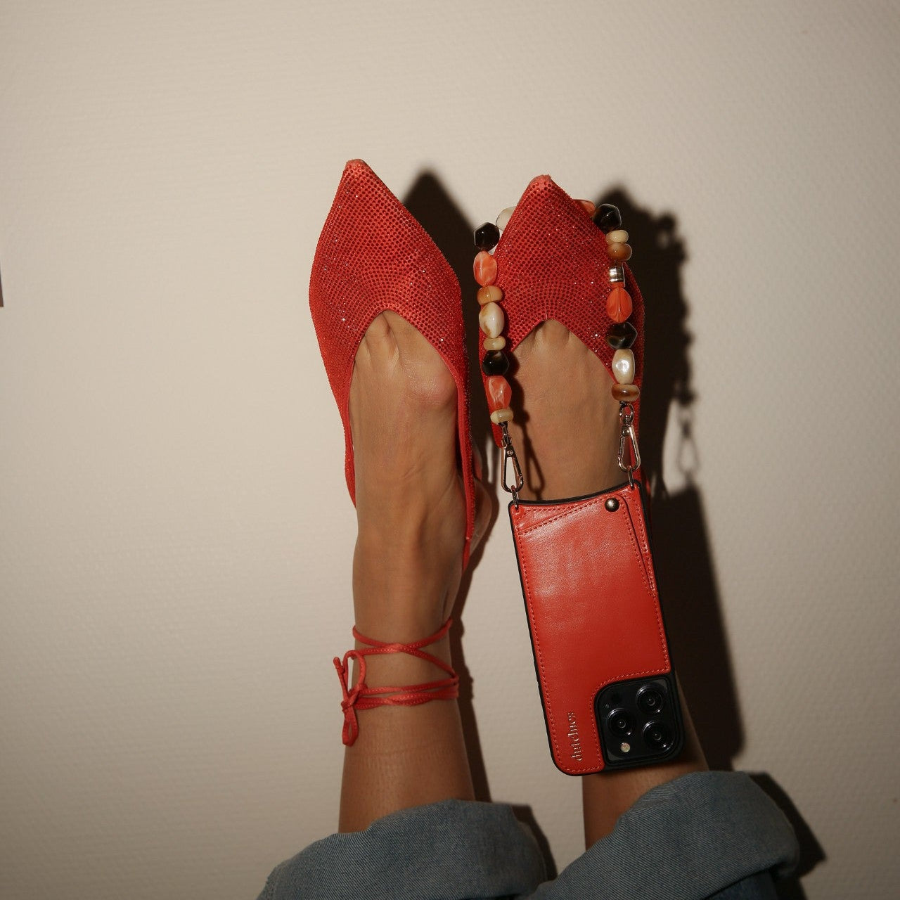 A stylish flat lay of red pointed shoes with a phone charm | beaded | Sonia attached to a sleek phone case.