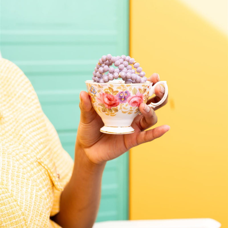 A person holding a delicate floral teacup filled with purple beads, set against a vibrant background.
