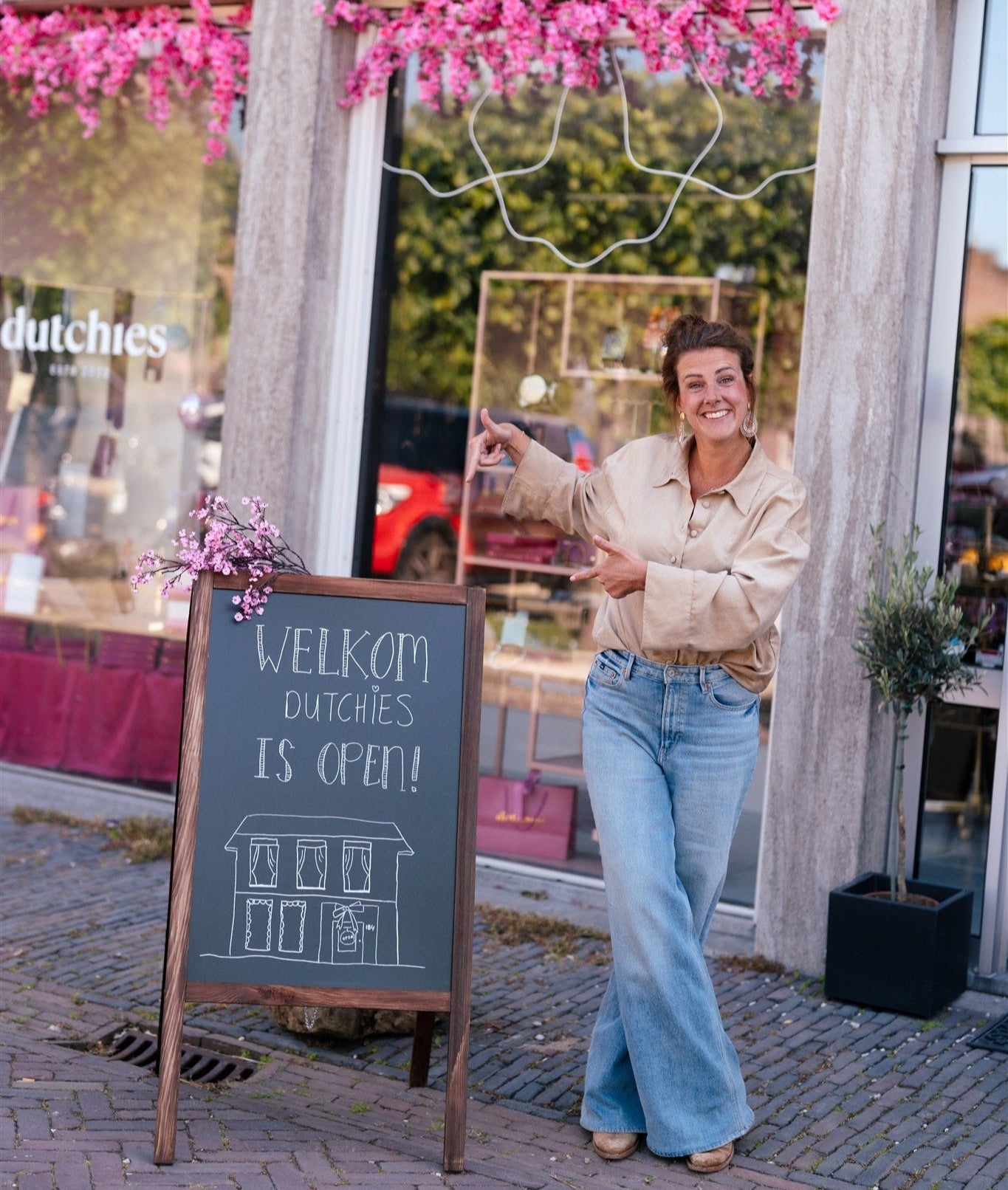 Woman standing next to a 'Welkom Dutchies is open!' sign outside a store with pink flowers and 'dutchies' branding.