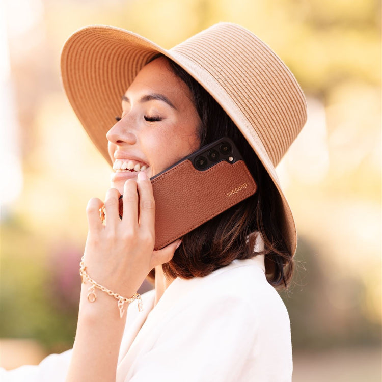 Woman wearing a straw hat, smiling while using her phone with a stylish case, showcasing a happy moment.