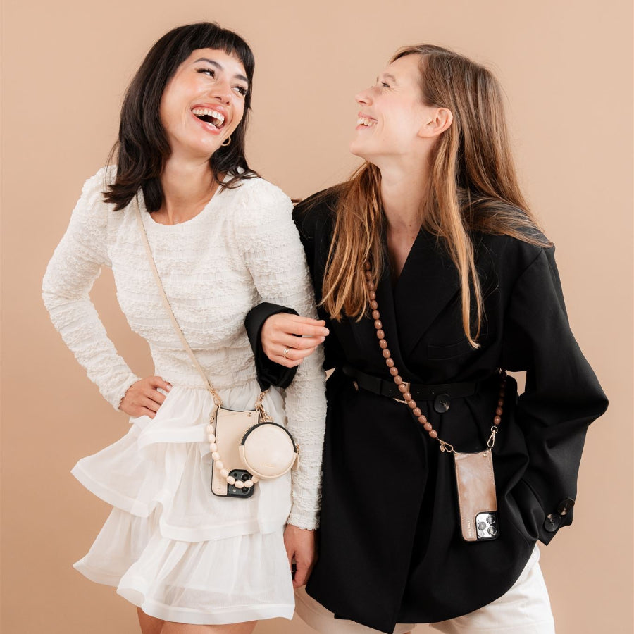 To kvinder iført perlebesatte mobilkæder og stilfulde outfits, smilende mod en neutral baggrund.
