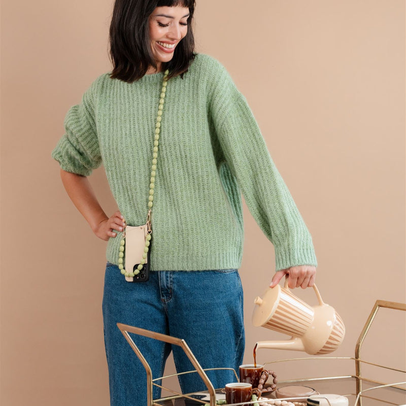Woman in a green sweater with beaded phone chain, smiling and holding a kettle, stylish home décor.