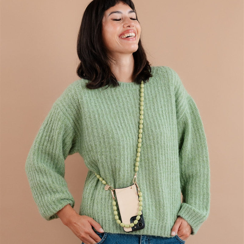 Woman wearing a green sweater and beaded phone chain, smiling against a beige background.