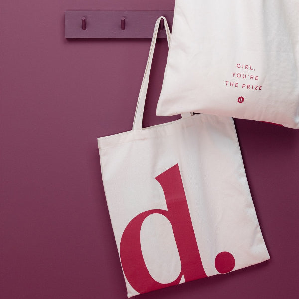 Totebag hanging on a wall hook, featuring a fun graphic design that empowers women.
