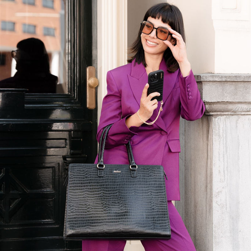 Stylish woman in purple suit holding a smartphone with the Laptop Bag Sharon | Noir at her side, showcasing fashionable elegance.