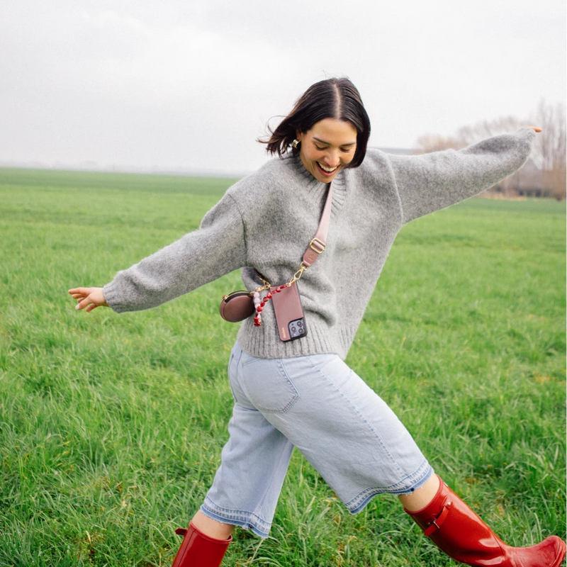 Woman in gray sweater joyfully showing off her Phone Strap | Basic | Mauve, styled with casual denim in a green field.