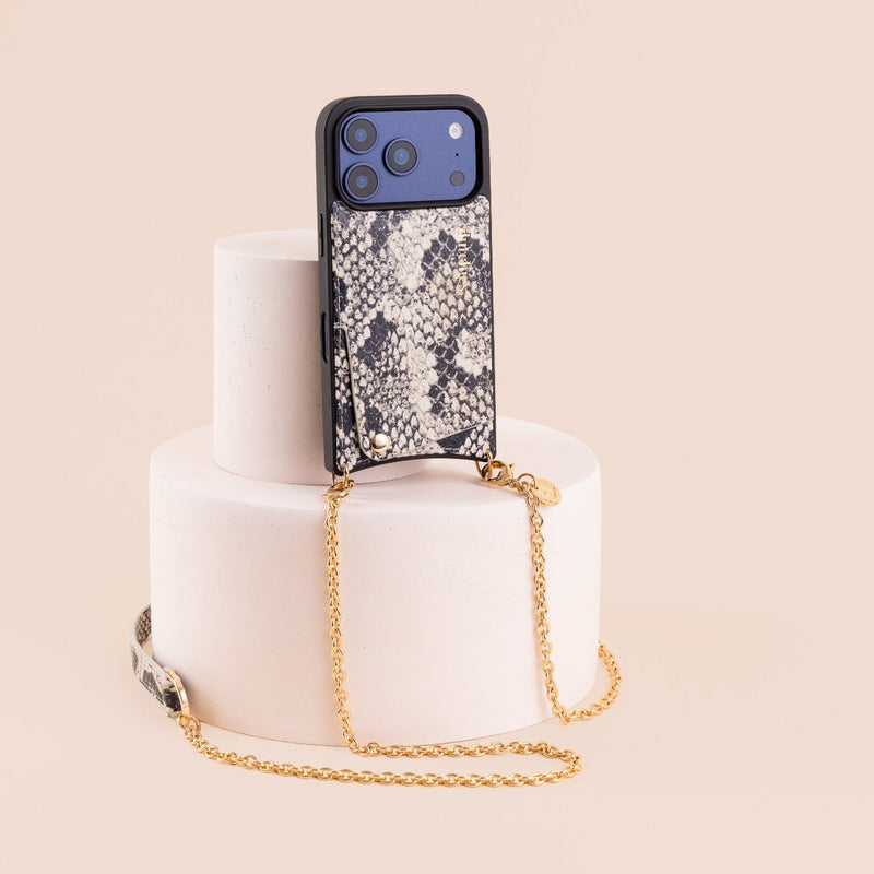 A smartphone case with a black and beige snakeskin pattern, featuring a gold chain strap and a black camera bezel, displayed on a white tiered pedestal against a light beige background.