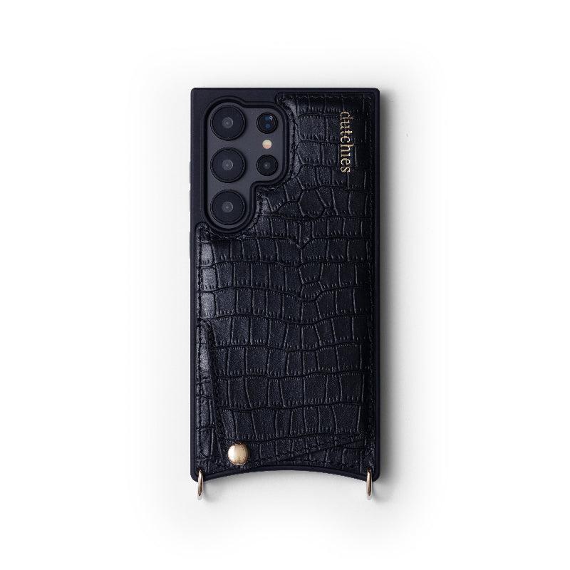 A black leather mobile phone case with gold metal finishes and a crocodile pattern. It features a camera cutout, a brand logo, and two rings presumably for attaching a strap.