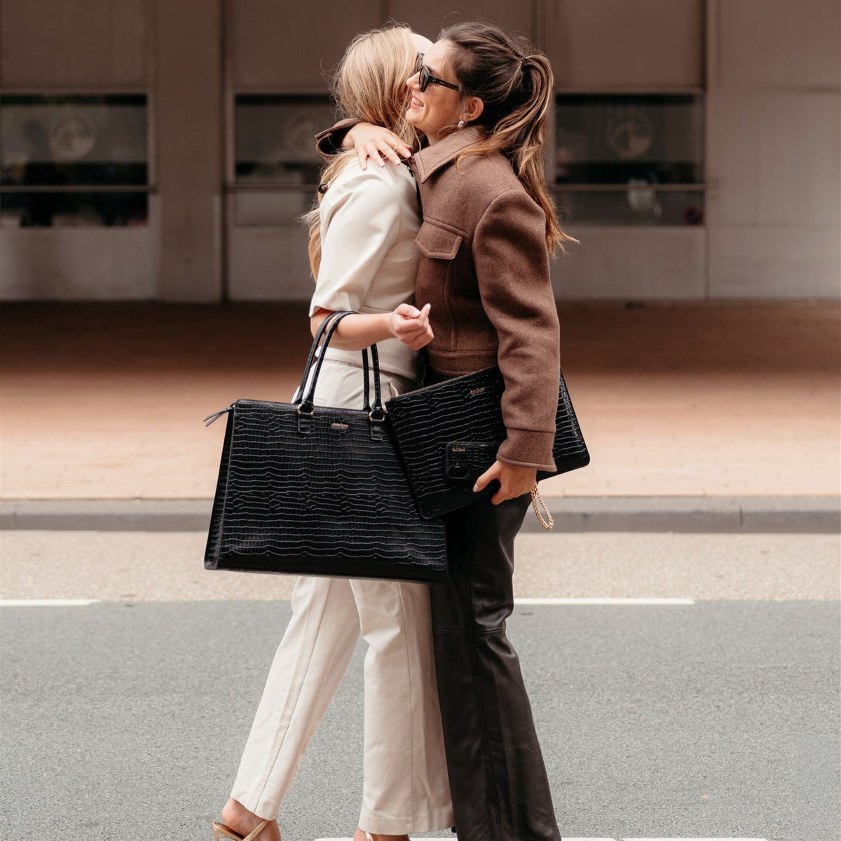 Deux femmes s’enlaçant avec joie tout en portant une élégante sacoche noire pour ordinateur portable, incarnant la mode urbaine moderne.
