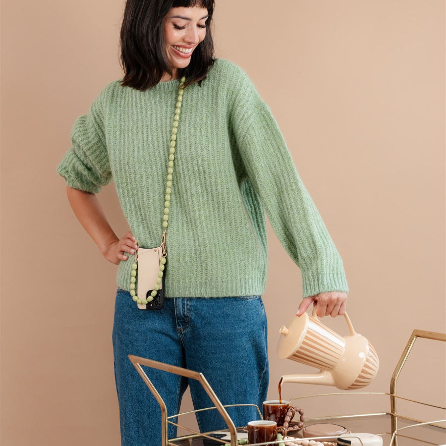 Woman in a green sweater with beaded phone chain, smiling and holding a kettle, stylish home décor.