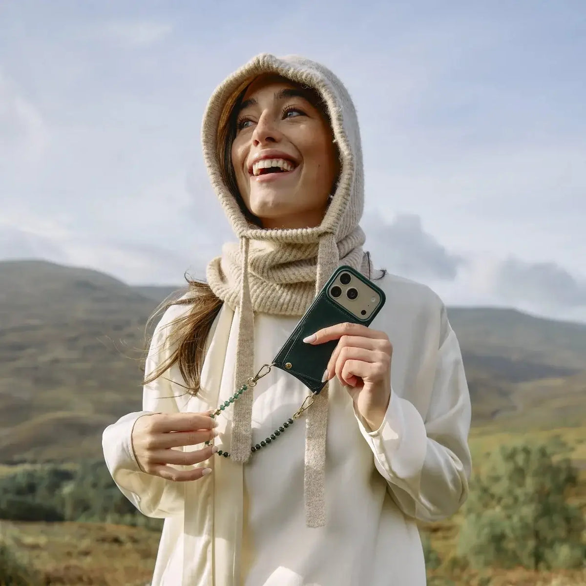 A joyful woman wearing a cozy scarf, holding her phone adorned with a Phone Charm | Beaded | Nina in a scenic outdoor setting.