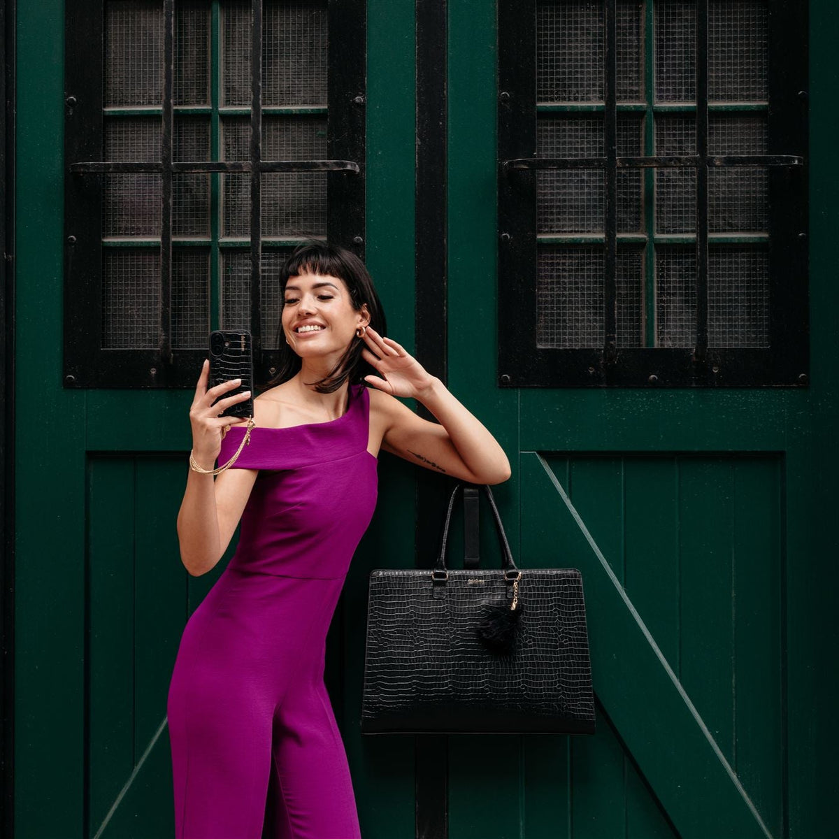 Femme en combinaison violette posant avec une sacoche en cuir noir pour ordinateur portable devant une porte verte.