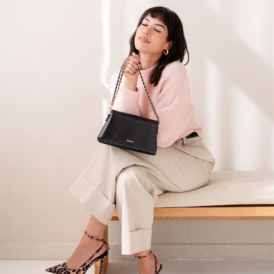 Sophie Crossbody Bag | Noir Gold holdt af en kvinde i en hyggelig pink sweater, der fremviser dens elegante stil og praktiske anvendelighed.