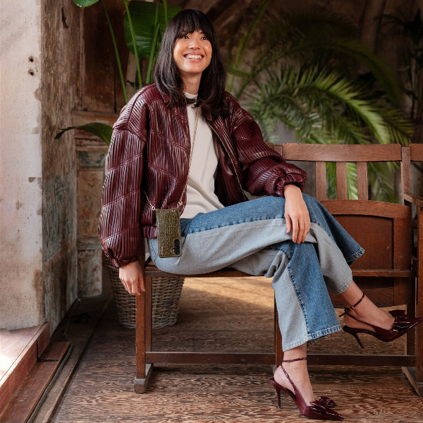 A stylish woman wearing a burgundy jacket, seated casually with olive green phone chain, embodying chic fashion.