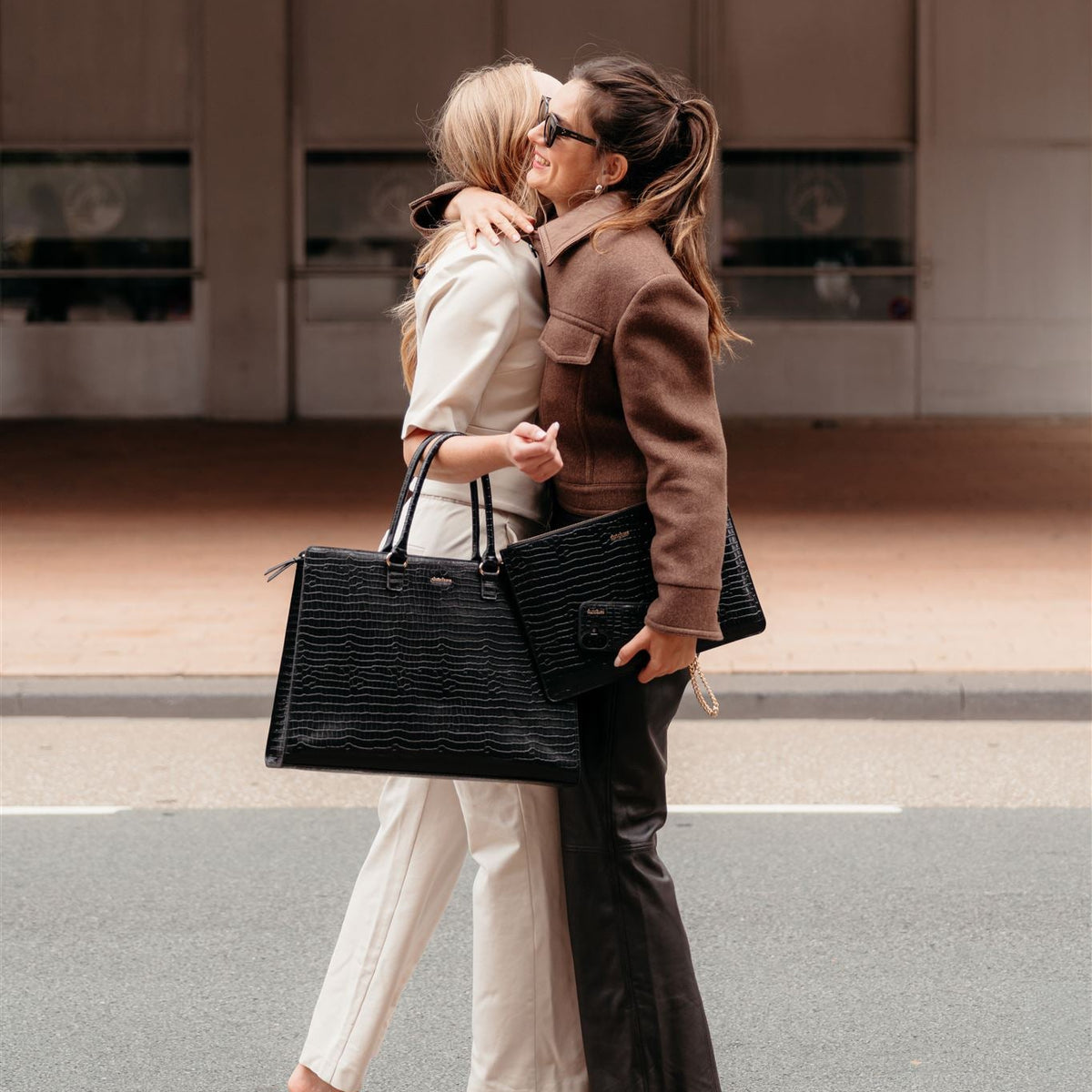 Zwei Frauen umarmen sich und präsentieren dabei stilvolle schwarze Laptoptaschen in einem schicken urbanen Setting.